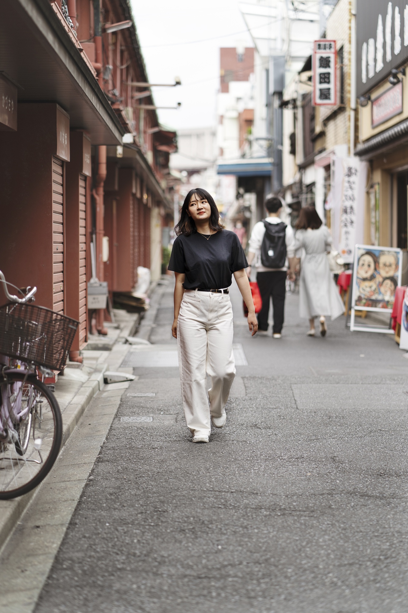 黒いTシャツ姿の女性が商店の並ぶ静かな通りを歩く様子